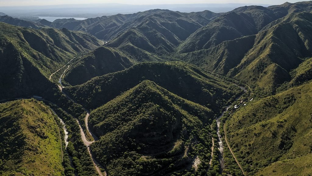 Vista aérea de la quebrada del Indio Bamba: dos cordones de sierra cubiertos de monte bajan hacia un valle en V, con el río corriendo por el fondo y el camino de montaña serpenteando entre las laderas. Al fondo, el lago y la llanura de Córdoba.